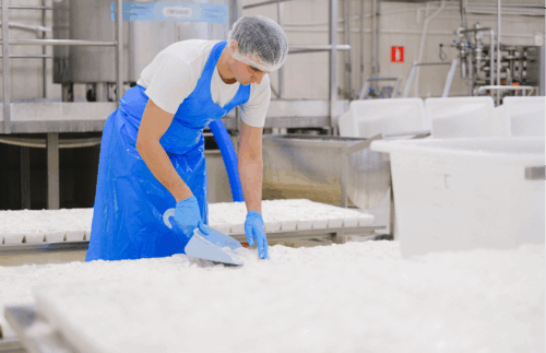 Arbeiter in Schutzkleidung mit Haarnetz, Schürze und Handschuhen schaufelt weißes Material in einer Produktionshalle.